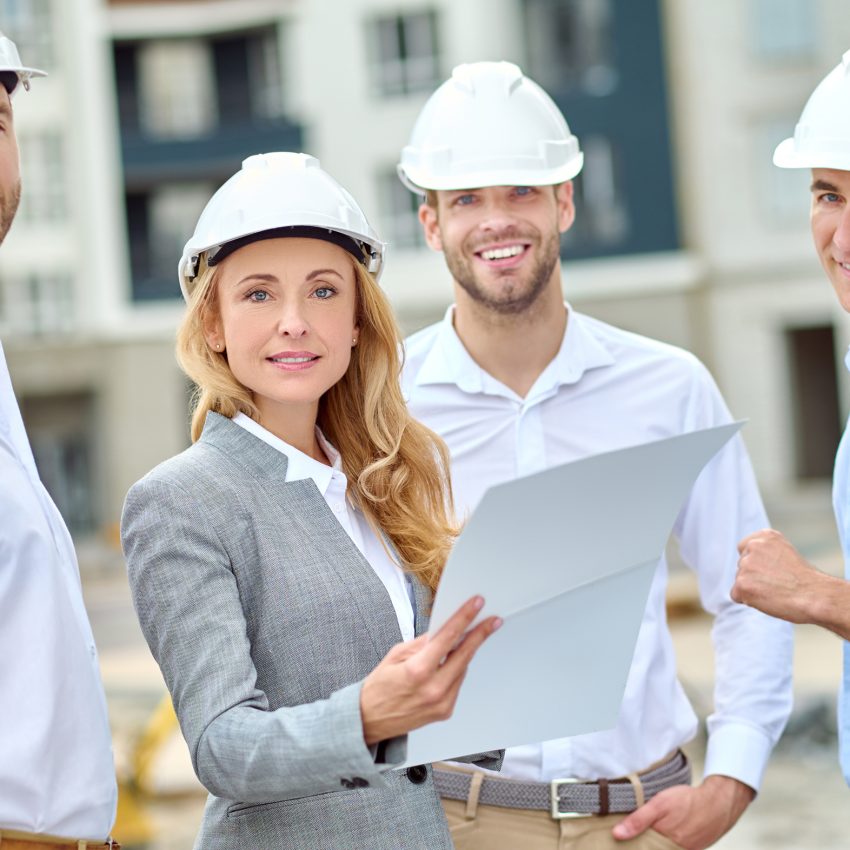 Waist-up portrait of a beautiful tranquil confident blonde female supervisor and construction workers in hardhats standing outdoors